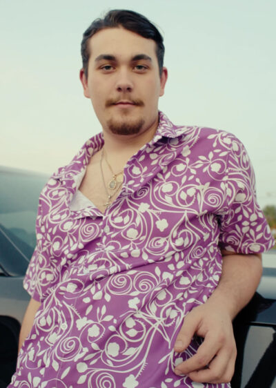 A man with dark hair and a beard leans against a car, confidently wearing the Biscuit Oliva Baki Button Up Hawaiian Shirt paired with gold necklaces, and looks directly at the camera.