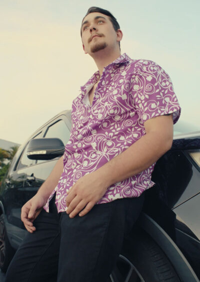 A man in a Biscuit Oliva Baki Button Up Hawaiian Shirt and black pants leans against a black car, gazing thoughtfully into the distance under a clear evening sky.