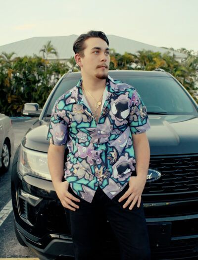 A man with short dark hair and facial hair stands in front of a black Ford SUV, wearing the Ghost Type Pokemon Pokémon Button Up Hawaiian Shirt. He has his hands on his hips, looking to the side with palm trees and a building behind him.