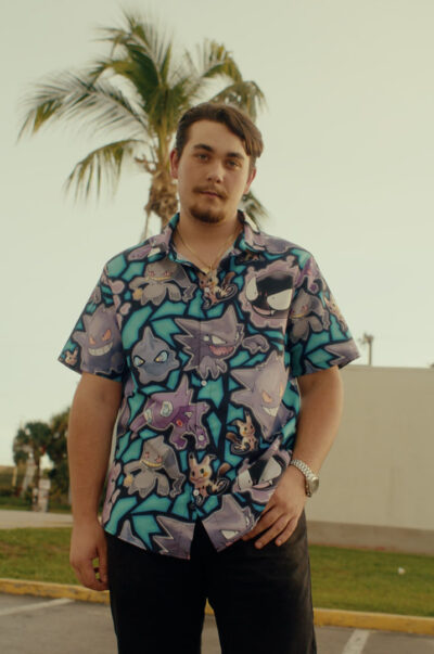 A young man stands outdoors near palm trees, wearing the Ghost Type Pokemon Pokemon Button Up Hawaiian Shirt and black pants, looking at the camera with a neutral expression.