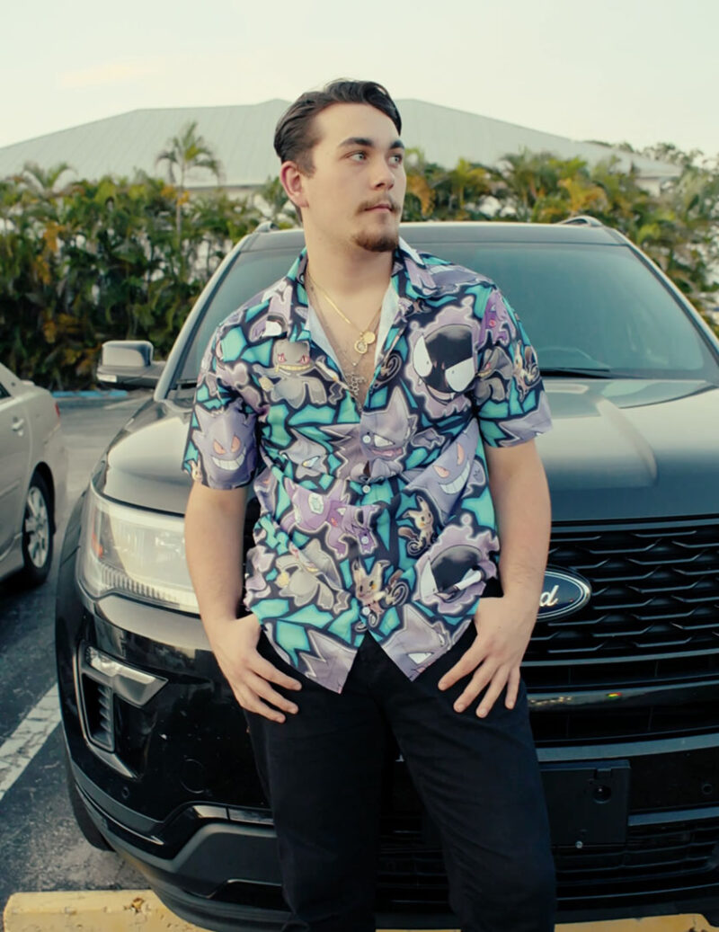 A young man with short hair and a goatee stands in front of a black Ford SUV, wearing the Ghost Type Pokemon Pokémon Button Up Hawaiian Shirt, black pants, and gold jewelry, with a building and trees in the background.