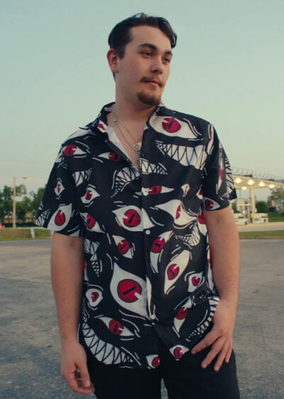 A man with short dark hair, a mustache, and a slight beard stands outdoors among trees and lit structures, wearing the Pride Fullmetal Alchemist Button Up Hawaiian Shirt—a black short-sleeve shirt featuring bold eye and teeth patterns.