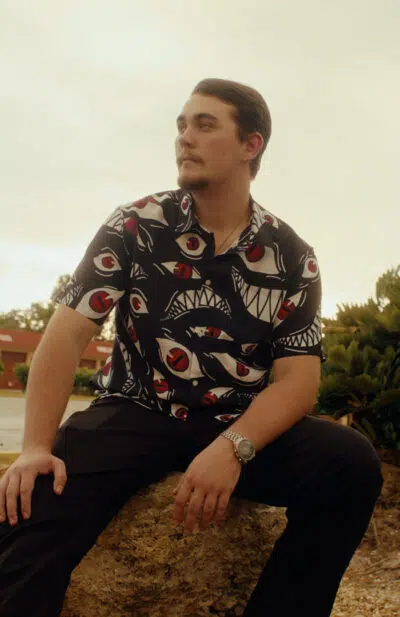 A man with dark hair and a mustache sits on a rock outdoors, wearing the Pride Fullmetal Alchemist Button Up Hawaiian Shirt and black pants, looking to the side against a warm, golden sky.