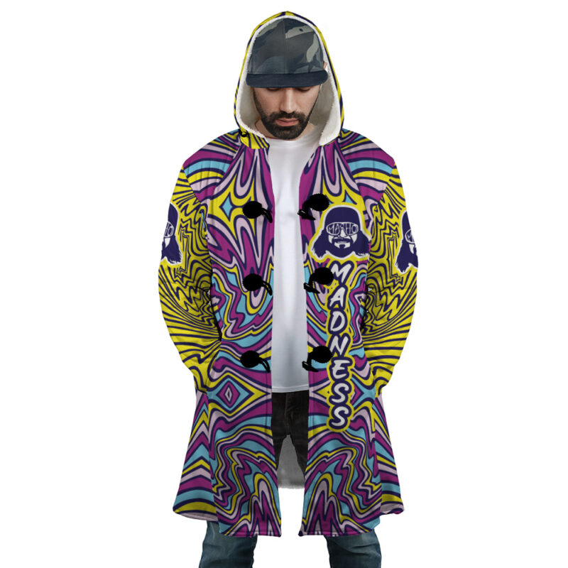 A man faces forward wearing the Trippy The Cream of the Crop Randy Savage Dream Cloak Coat, featuring bold psychedelic patterns in yellow, purple, pink, and blue, with MADNESS vertically printed and graphic logos on the upper sleeves.