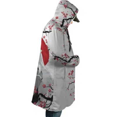 A person in profile with hands in pockets wears the Inuyasha Cherry Blossom Dream Cloak Coat, a hooded coat featuring cherry blossoms, a red sun, and a stylized white crane inspired by Japanese design.