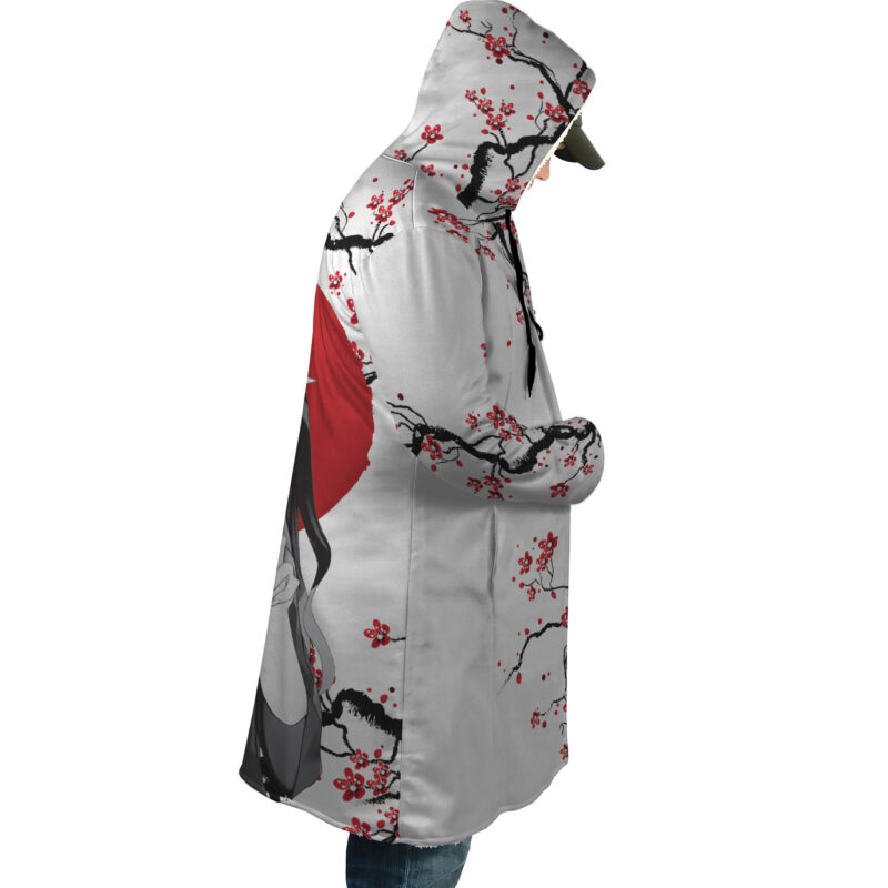 A person stands in profile with hands in pockets, wearing the Nezuko Kamado Cherry Blossom Demon Slayer Dream Cloak Coat featuring red cherry blossoms, black branches, and a large red circle on an artistic hooded design.