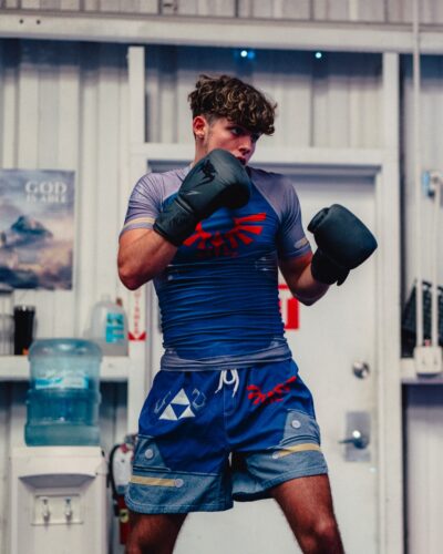A young man with curly hair wears the Zelda Shield Style Zelda Short Sleeve Rash Guard Compression Shirt and boxing gloves, standing in a fighting stance indoors. A water cooler and poster appear in the background.