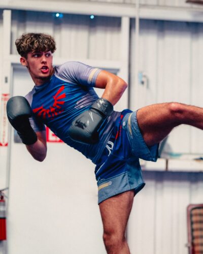 A young man in athletic gear and boxing gloves performs a high kick in the gym, wearing the Zelda Shield Style Zelda Short Sleeve Rash Guard Compression Shirt and blue shorts, looking focused and determined.
