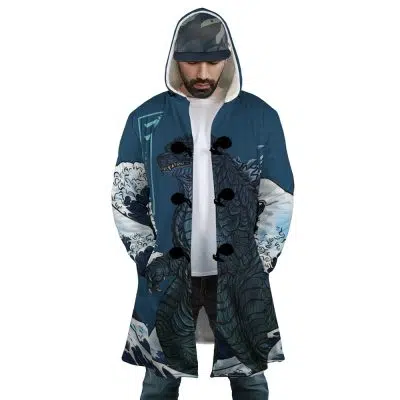 A man wears the Godzilla Dream Cloak Coat, a long blue hooded coat featuring wave graphics and a large sea creature inspired by Japanese art. He pairs it with a cap, looks down, and has his hands in his pockets.