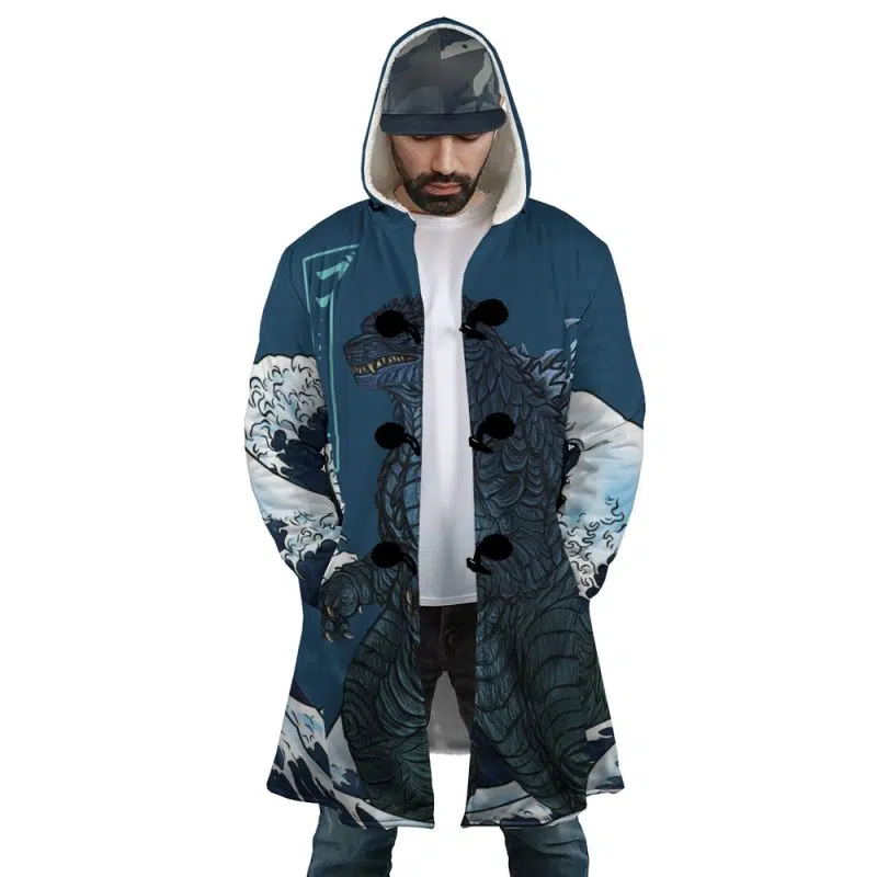 A man wears the Godzilla Dream Cloak Coat, a long blue hooded coat featuring wave graphics and a large sea creature inspired by Japanese art. He pairs it with a cap, looks down, and has his hands in his pockets.