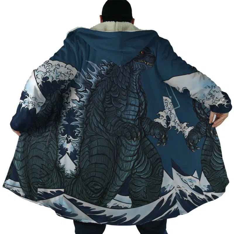 A person wearing the Godzilla Dream Cloak Coat, seen from behind with arms outstretched, showcases a large, detailed Godzilla illustration among ocean waves on the back of the long blue coat.