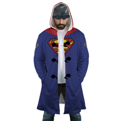 A man wearing the Superman DC Comics Dream Cloak Coat, featuring a long blue design with a red Superman logo on the chest and shoulders, a red hood, black toggles, plus jeans and a cap. He looks down with his hands in his pockets.