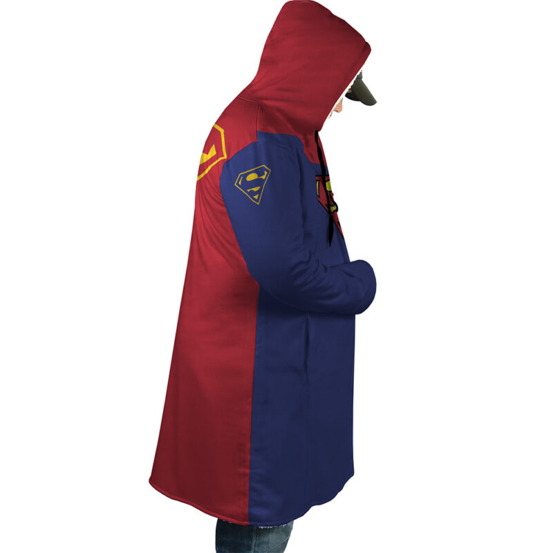 A person wears the Superman DC Comics Dream Cloak Coat, a split-color hooded coat—half red, half blue—with superhero logos on the back and sleeve. They stand sideways, facing right, hands in pockets, hood up.