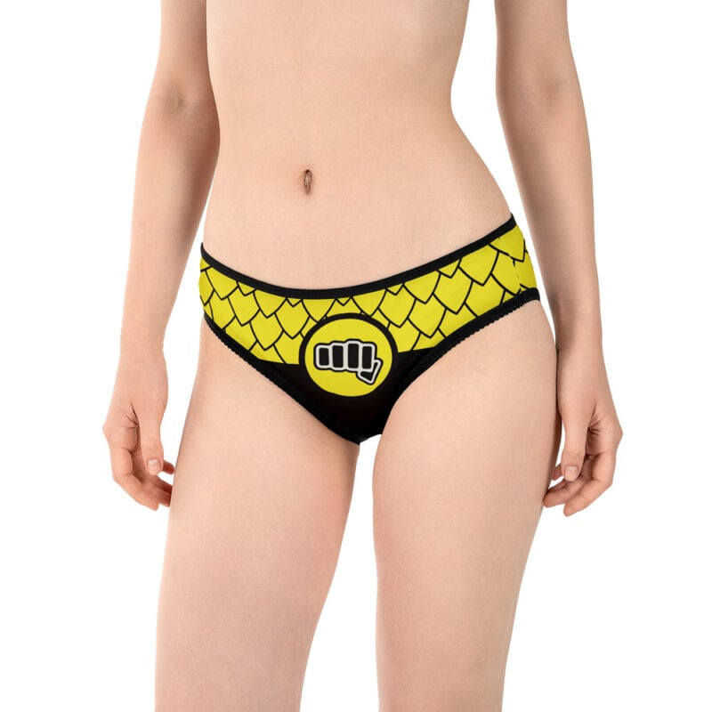 A woman models Cobra Kai Panties featuring a black and yellow scale pattern and a fist symbol on the front. She stands against a white background, with her face out of frame, showing only her upper body and legs.