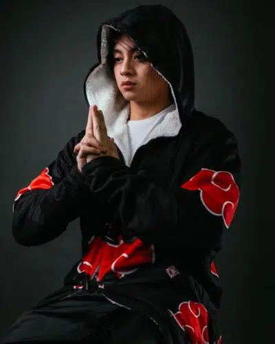 A person wearing a black hooded cloak with red cloud patterns poses with their hands together in a ninja hand sign against a dark background.