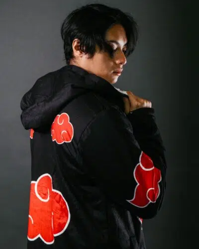 A person with short dark hair wears a black hoodie decorated with red clouds, looking over their shoulder against a dark background.