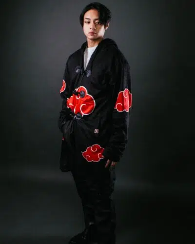 A person stands against a dark background wearing a black cloak decorated with large red clouds outlined in white, resembling the Akatsuki cloak from the anime Naruto.