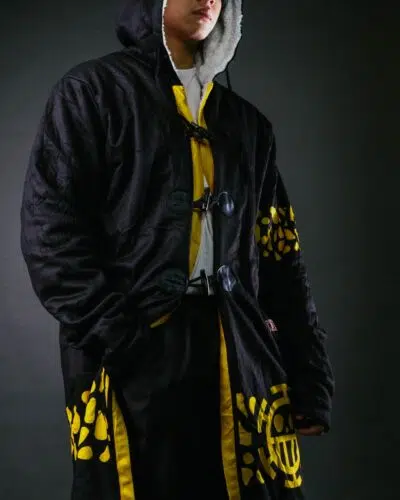 A person wearing a black hooded coat with yellow geometric patterns, white inner lining, and large toggle buttons, paired with matching black and yellow pants, standing against a dark background.
