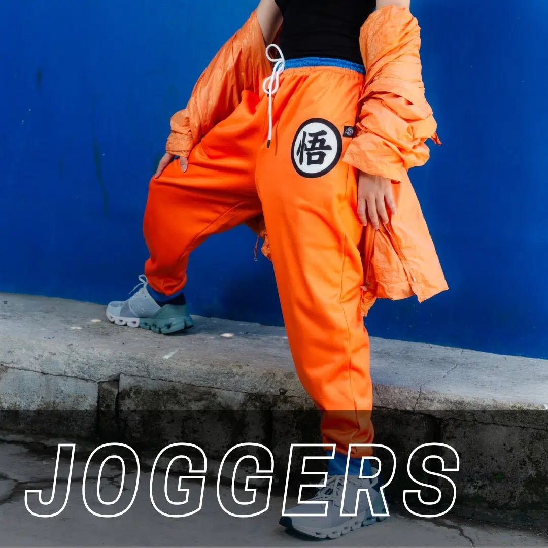 A person poses against a blue wall, wearing bright orange jogger pants with a black and white patch and matching jacket, paired with gray sneakers. The word JOGGERS is overlaid at the bottom in large, outlined letters.