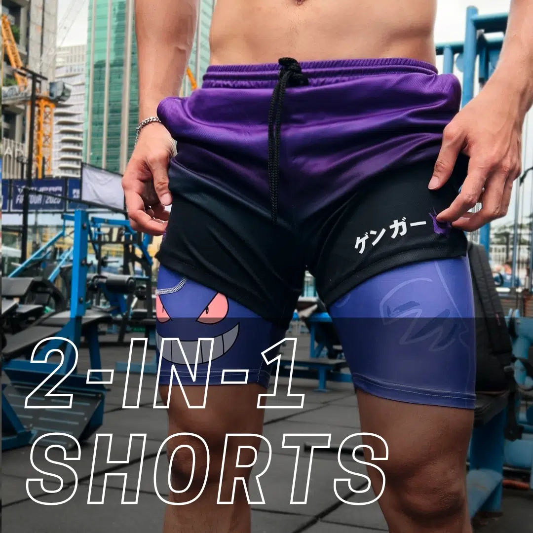A person wearing purple and black 2-in-1 athletic shorts featuring a cartoon face and Japanese characters stands in an outdoor gym with city buildings in the background. Text on the image reads “2-IN-1 SHORTS.”.