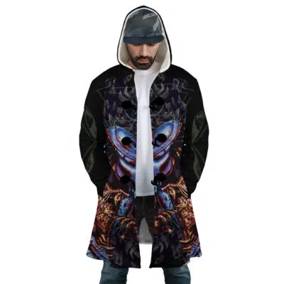 A man wears the Crusader Templar Cross Dream Cloak Coat, featuring a bold abstract pattern. He stands with hands in pockets, paired with a cap and jeans, looking down with a neutral expression.