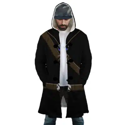 A man wearing the Uchiha Successors Shippuden Dream Cloak Coat—a long, hooded black coat with brown straps and silver buckles—over a white shirt, blue jeans, and a patterned cap. He has a beard and looks down.