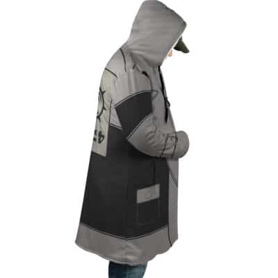 Wearing the Sidon The Legend of Zelda Dream Cloak Coat, a person stands in profile with hands in pockets, looking down. The gray and black hooded coat features graphic designs paired with a green cap.