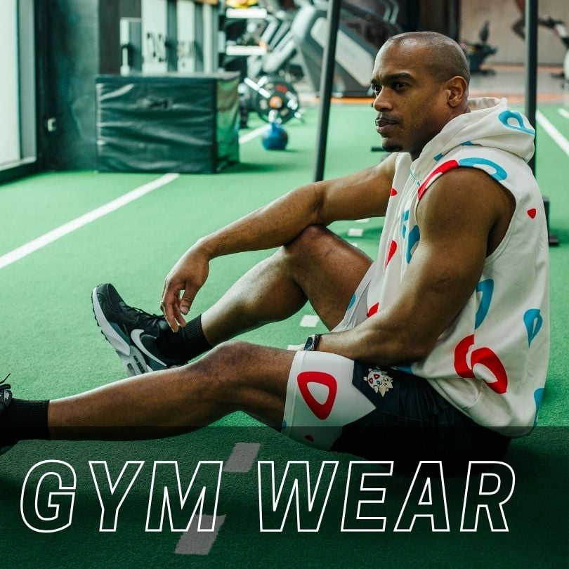 A man in a sleeveless patterned hoodie and athletic shorts sitting on a green gym floor with the text GYM WEAR overlaid at the bottom.