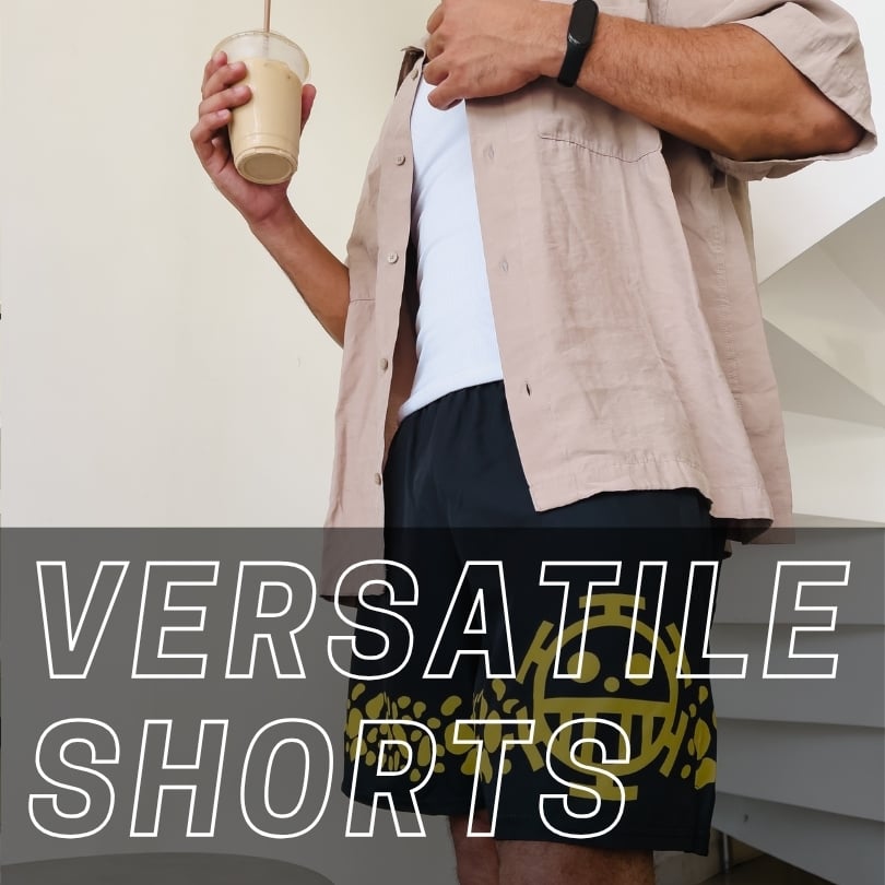 A person wearing a tan button-down shirt over a white tank top and black shorts with a yellow graphic design, holding an iced coffee. Text overlay reads: VERSATILE SHORTS.